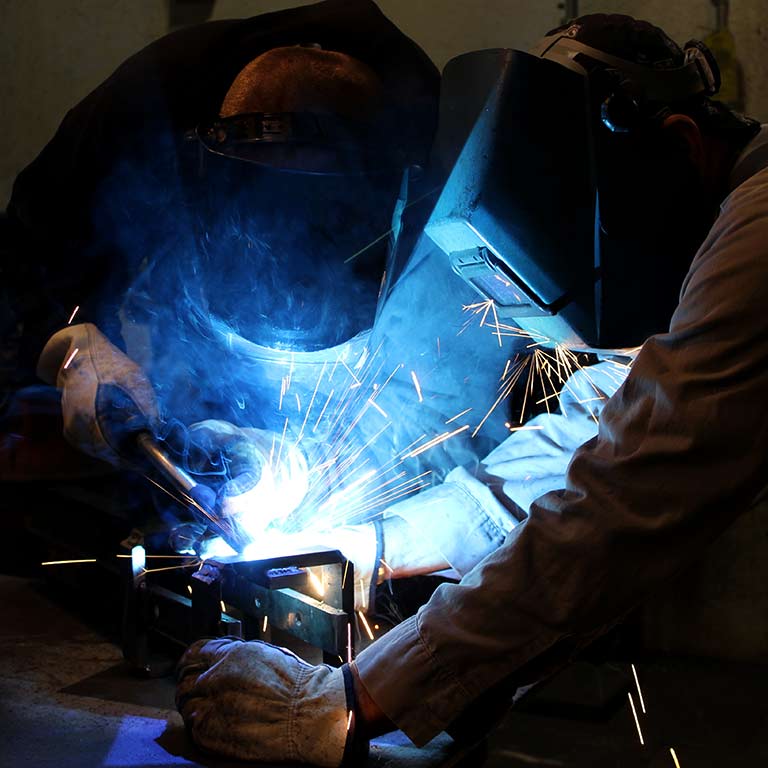 Two people wear welding masks and operate a blowtorch.
