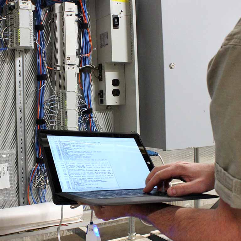 A person holds a laptop in front of a cabinet filled with wiring and components.