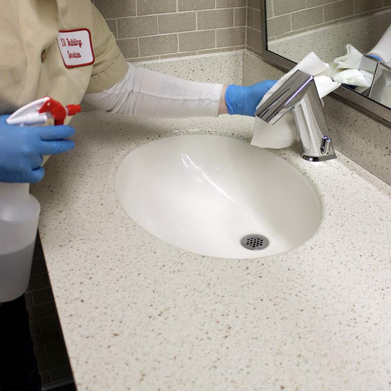 A custodial worker with a spray bottle and paper towel wipes down a sink.